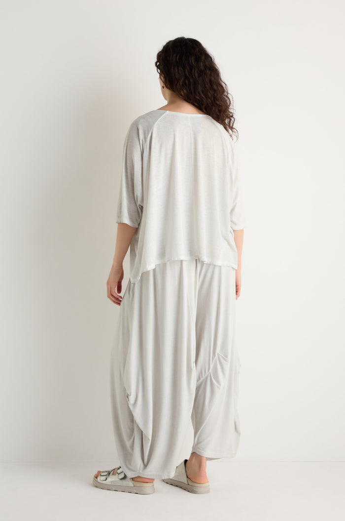 A person with long curly hair, wearing the Seam Detail Jersey Top in a relaxed silhouette, paired with loose light gray clothing and sandals, stands facing a white wall with their back to the camera.