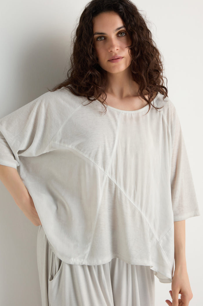 A woman with wavy brown hair stands against a white wall, looking at the camera, wearing the light beige Seam Detail Jersey Top in a relaxed fit with matching pants and subtle asymmetric seam detailing.
