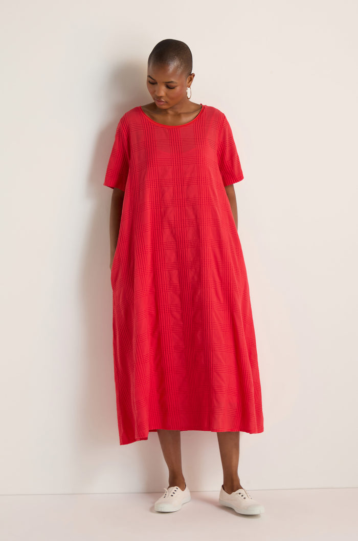 A person stands against a white wall wearing a loose-fitting, short-sleeved Self Check Flared Dress in red and white shoes.