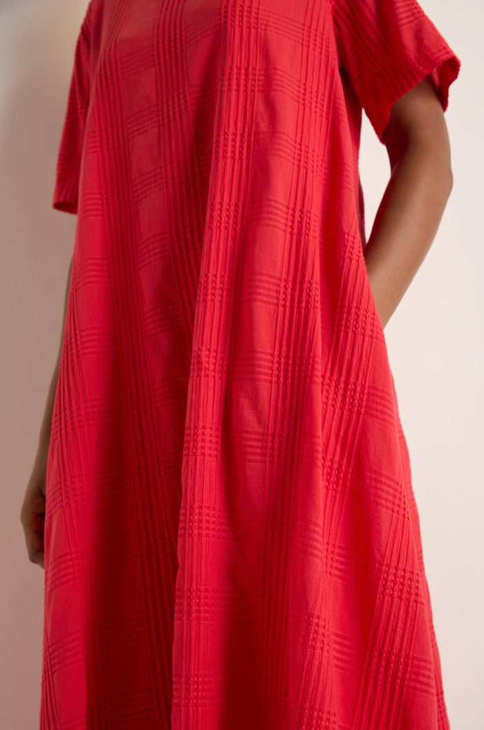 A person wears the Self Check Flared Dress—a loose-fitting, textured red cotton dress with short sleeves—standing against a plain light background.