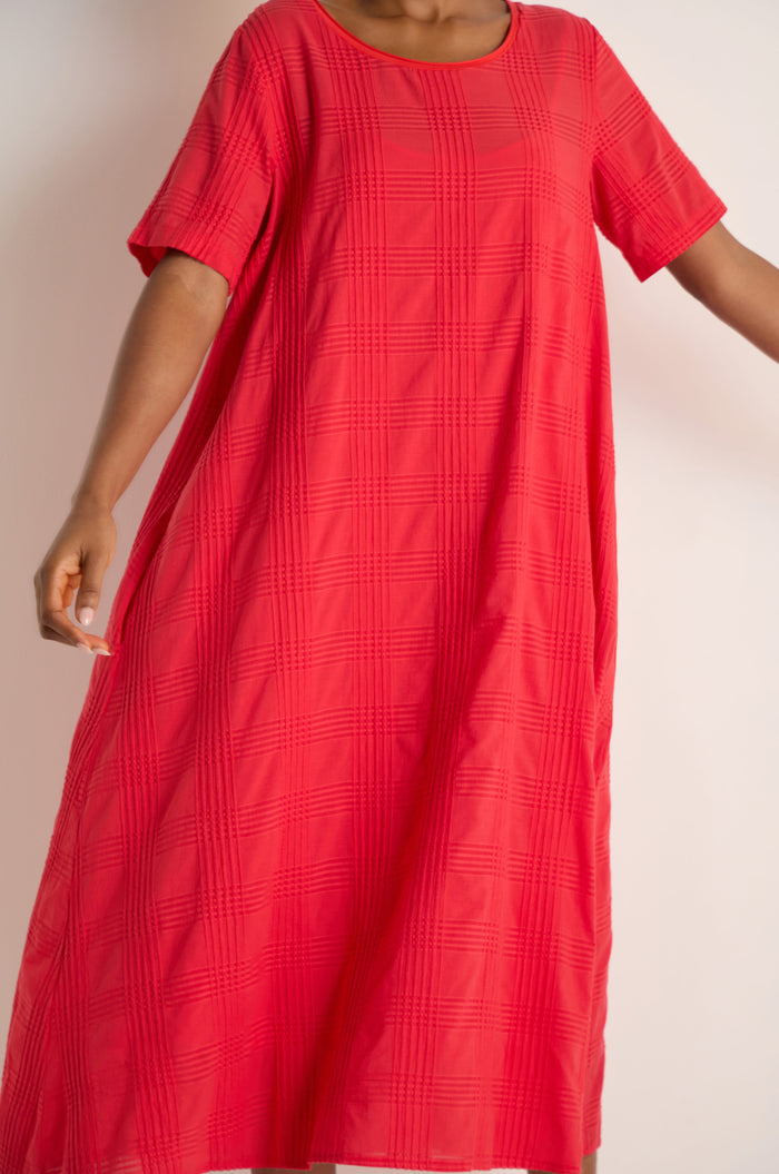 A person is shown from the torso up wearing a loose-fitting, short-sleeved Self Check Flared Dress in textured red cotton against a plain background.