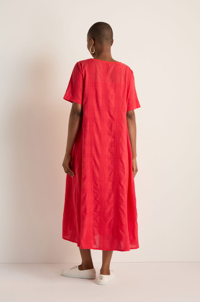 Person stands with back to camera, wearing the Self Check Flared Dress in textured red cotton and white sneakers, set against a plain light background.