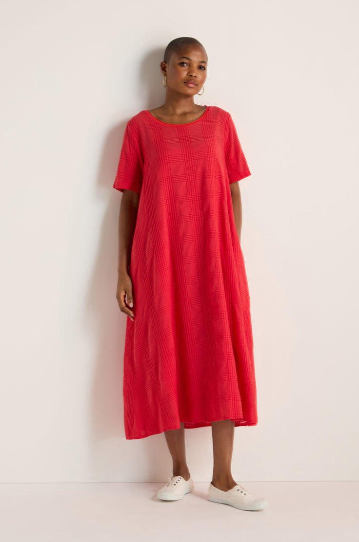A person stands against a plain white wall wearing the Self Check Flared Dress, a loose-fitting, bright red, textured cotton A-line midi dress, paired with white sneakers.