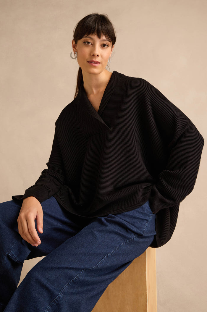 A woman with straight dark hair and hoop earrings is seated on a wooden stool against a beige background, wearing the Shawl Collar Black Ribbed Knit sweater with a relaxed fit and blue jeans.