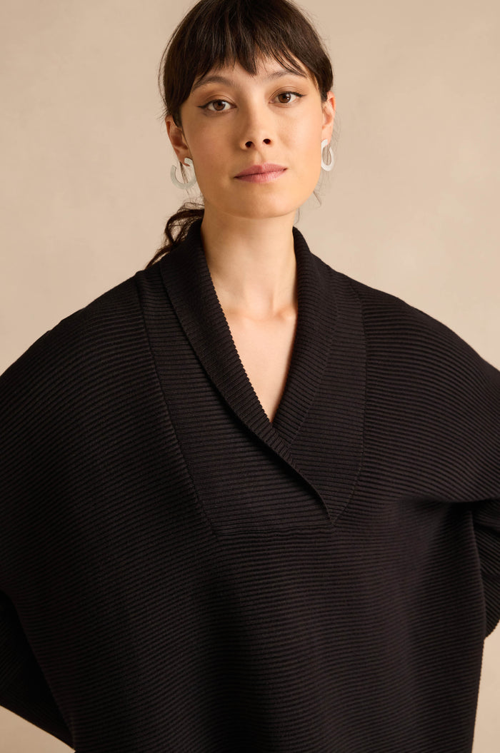 A woman with dark hair and bangs, wearing silver earrings and the Shawl Collar Black Ribbed Knit sweater with a relaxed fit, stands against a beige background.