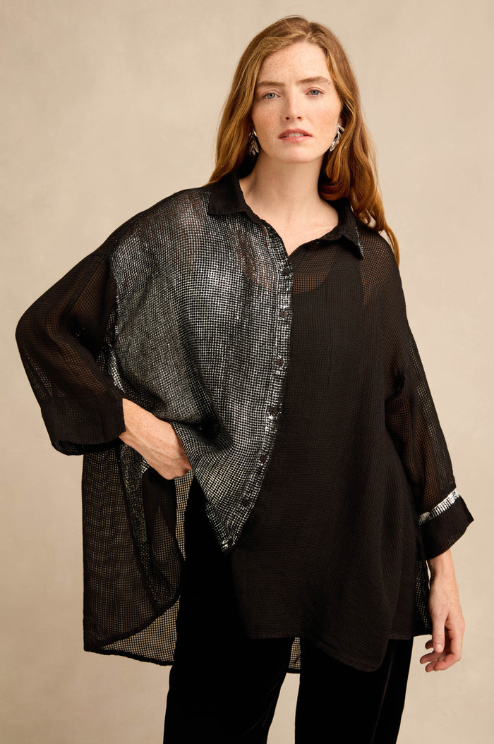 A woman stands against a neutral background wearing the Sheer Silver Panel Flared Shirt—a semi-sheer black, silk-blend top with a striking metallic silver panel—paired with black pants and silver earrings.