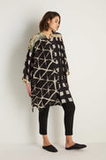 A person stands against a white wall wearing a relaxed-fit Shibori Print Long Shirt in black and beige, paired with black pants and black slip-on shoes.