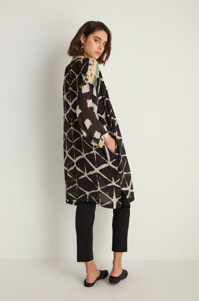 A person stands sideways with one hand in their pocket, wearing a relaxed fit, black and beige Shibori Print Long Shirt over black pants against a plain light background.