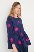 A woman wears the Shibori Spot Silk Shirt, a loose-fitting navy silk top with long sleeves and bright pink circular Shibori patterns, standing against a plain light background.