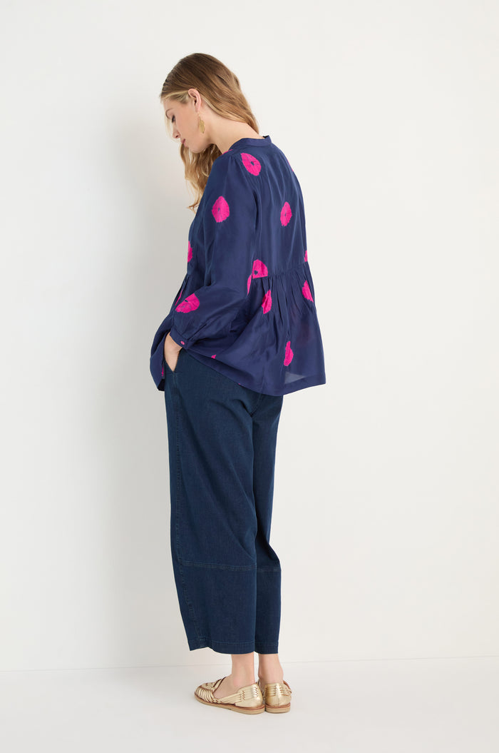 A woman stands in profile against a white background wearing the Shibori Spot Silk Shirt in navy with pink patterns, paired with wide-leg dark jeans and gold flats.