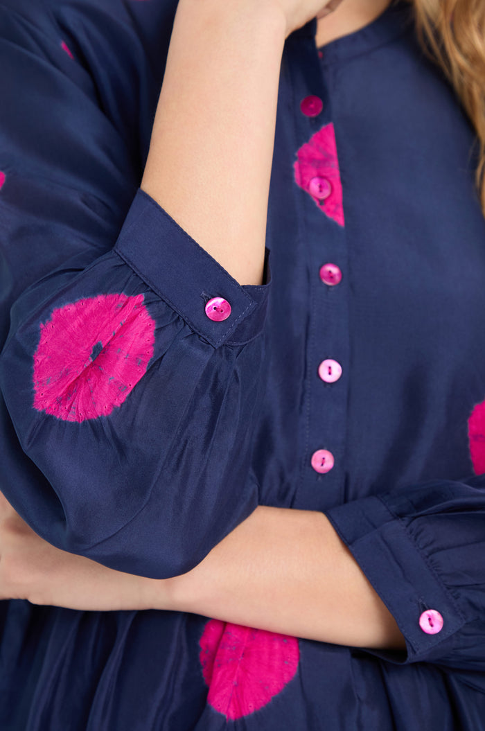 A person wears the Shibori Spot Silk Shirt, a navy blue silk top with bright pink circular patterns and matching pink buttons, inspired by Shibori design; one arm is crossed over their body.