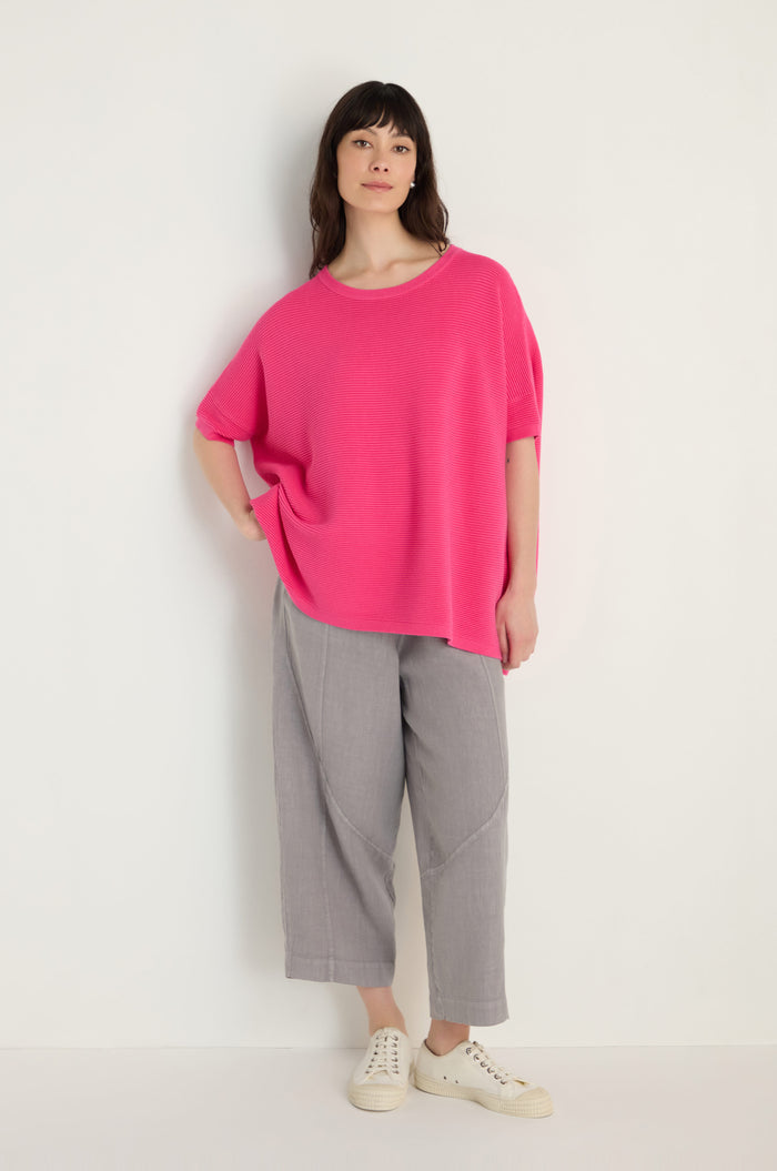A woman stands against a white wall wearing a Short Sleeve Hot Pink Ribbed Top, gray cropped pants, and white sneakers.