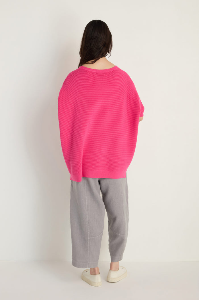 A person stands facing a white wall, dressed in a Short Sleeve Hot Pink Ribbed Top, loose gray pants, and white slip-on shoes.