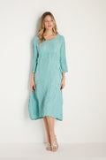 Woman stands against a white wall, wearing a loose-fitting Silk Linen Mint Patch Pocket Dress with three-quarter sleeves in light blue and white strappy sandals.