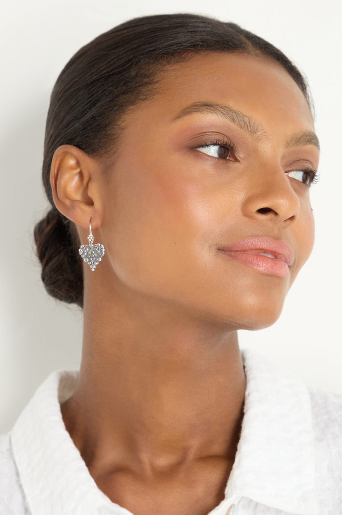 A woman with smooth dark hair in a bun wears a white textured top and Silver Heart Earrings, gazing slightly to the side against a plain background.
