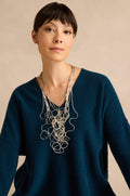 Woman with short dark hair wearing a blue V-neck sweater and the Silver Long Medley Necklace, posed in front of a beige background.