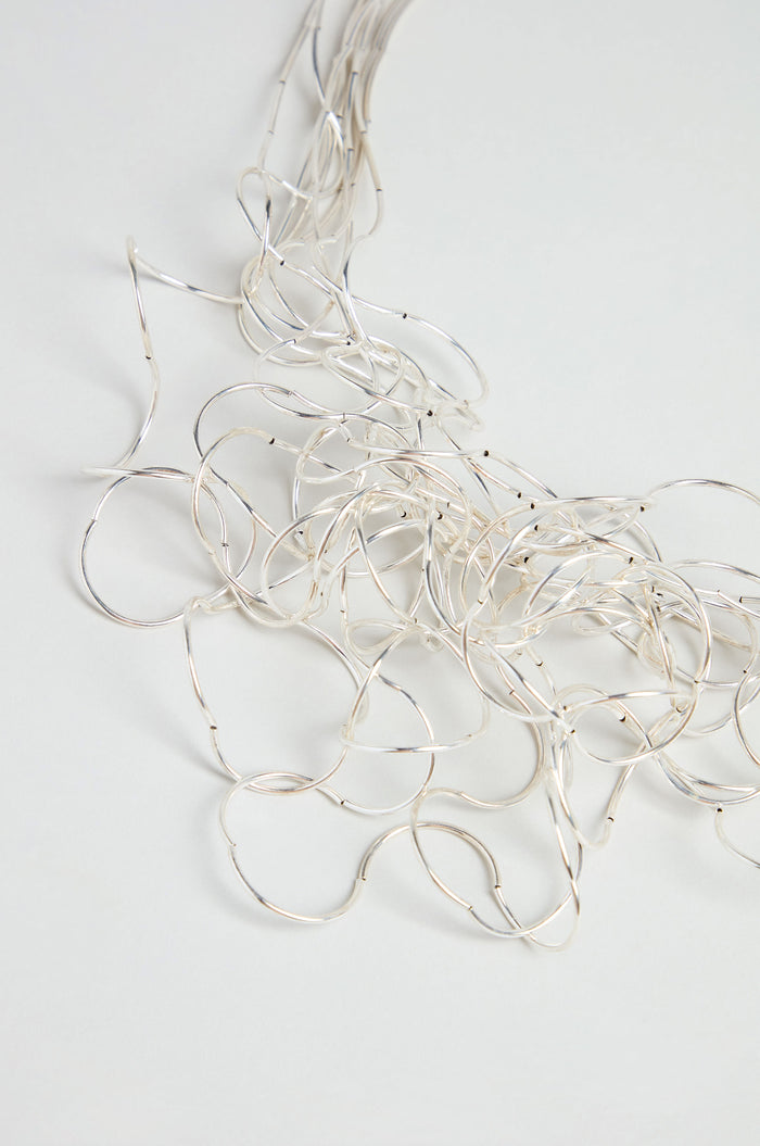 A Silver Long Medley Necklace, with its delicate strands of thin silver wires, is spread out on a light gray surface.