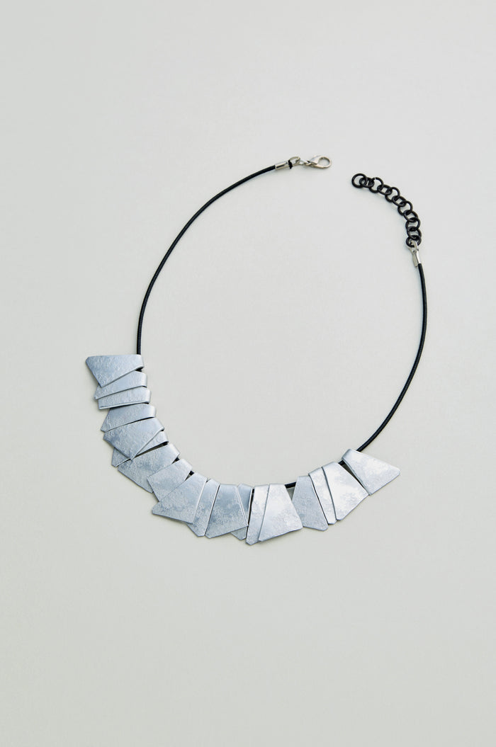The Mini Segments Necklace is a geometric accessory with angular, anodised aluminium segments on a black cord and adjustable clasp, showcased on a light gray background.