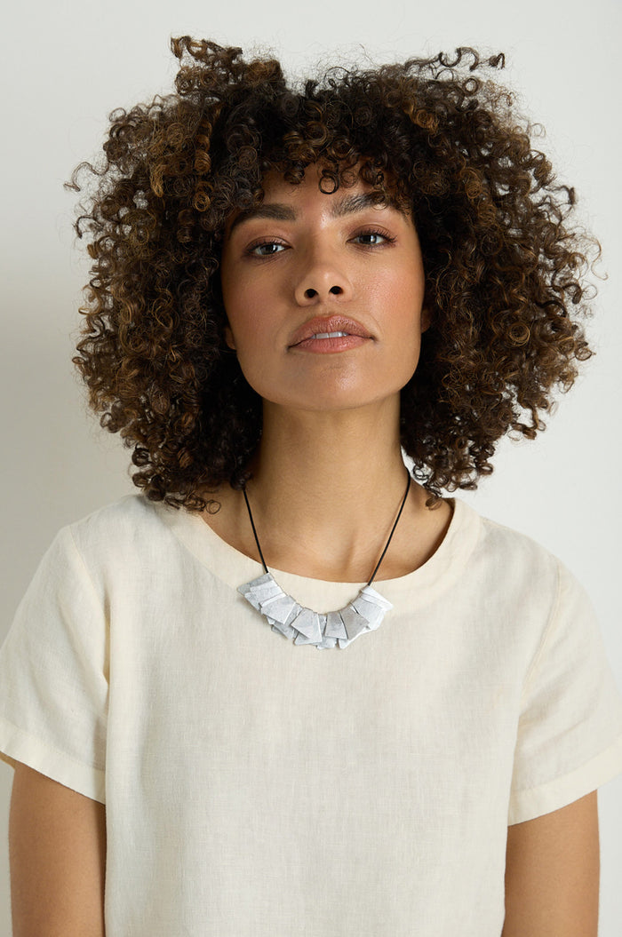 A woman with curly hair wears a cream linen top and the Mini Segments Necklace, a bold geometric jewelry piece, standing against a plain light background.