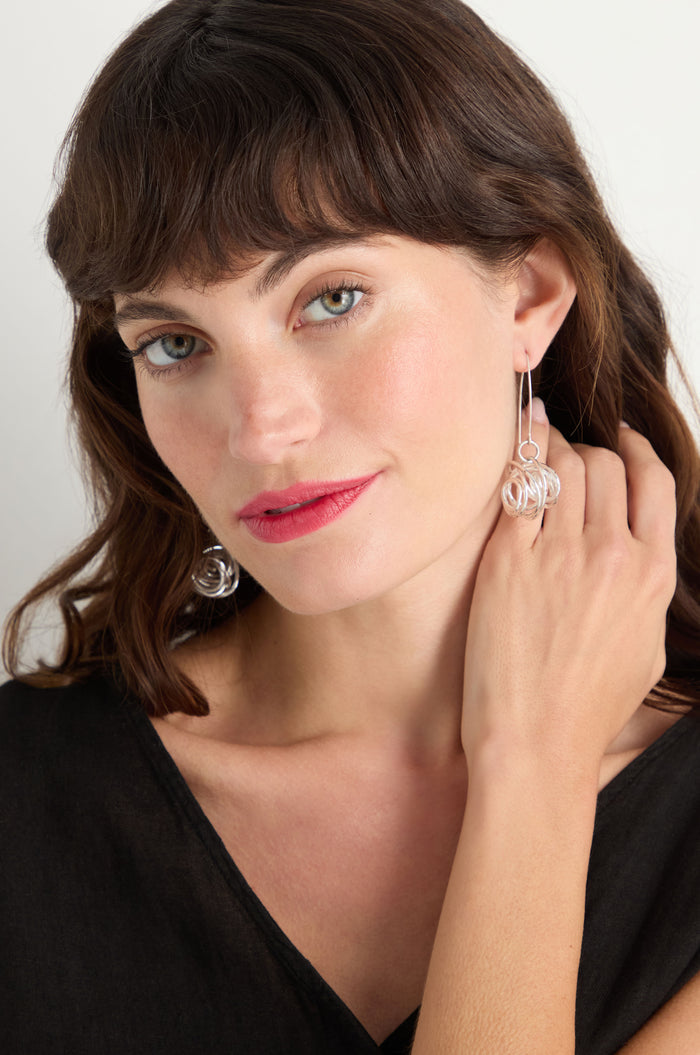 A woman with wavy brown hair and a black top touches her neck and looks at the camera, showcasing Silver Scribble Earrings.