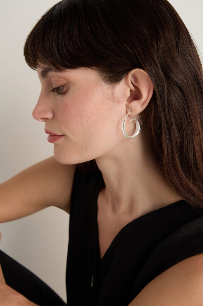 A woman with long brown hair wears the Silver Swirl Earrings, styled with a sleeveless black top and photographed in profile against a plain background.