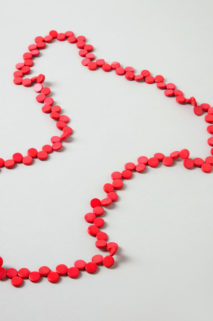 The Single Strand Coco Necklace, a long handcrafted piece, is arranged in loose wavy loops on a flat, light-colored surface.