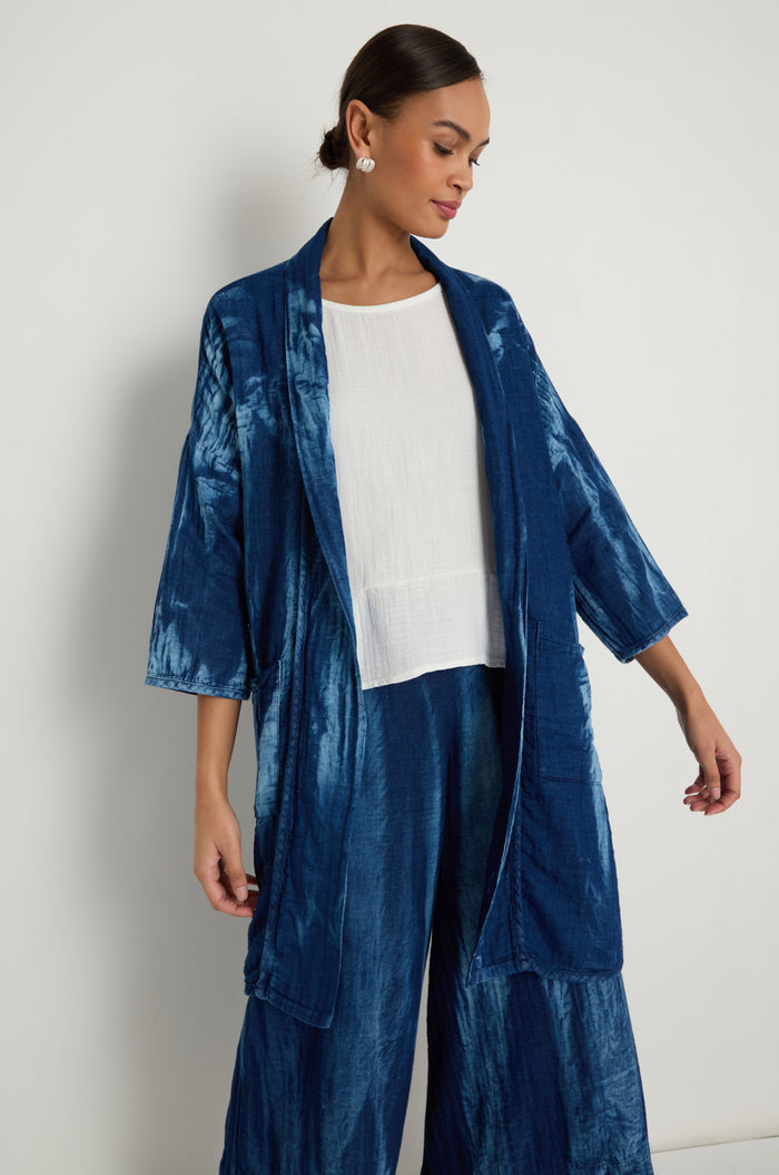 A woman stands against a plain white wall, wearing the Soft Denim Easy Jacket in a relaxed blue tie-dye style over a white top, paired with matching pants.