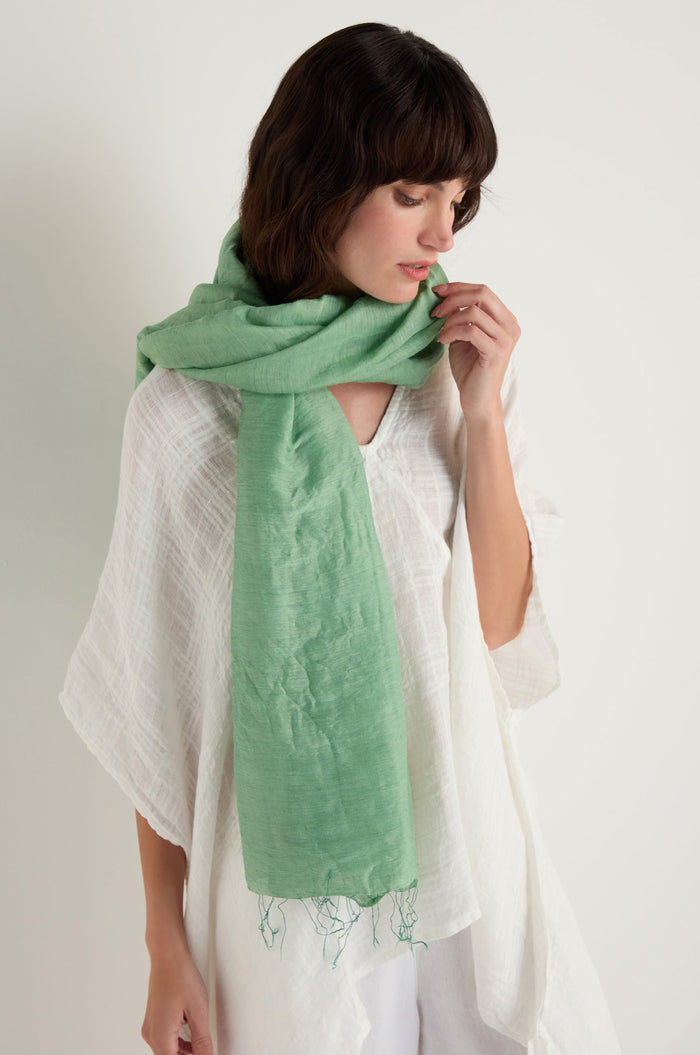 A woman in a white, loose-fitting top and pants wears the Olive Soft Linen Fringe Scarf around her neck, looking down against a plain white background.