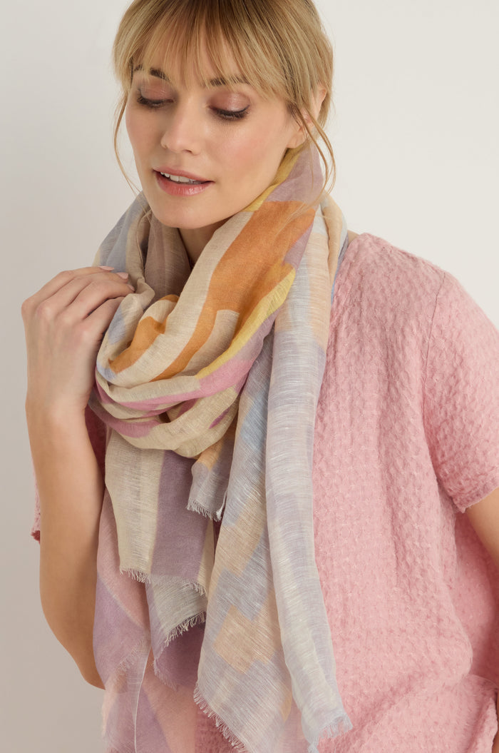 A woman wearing a textured pink top and the Soft Stripe Linen Blend Scarf stands against a plain light background, looking downwards with a neutral expression.