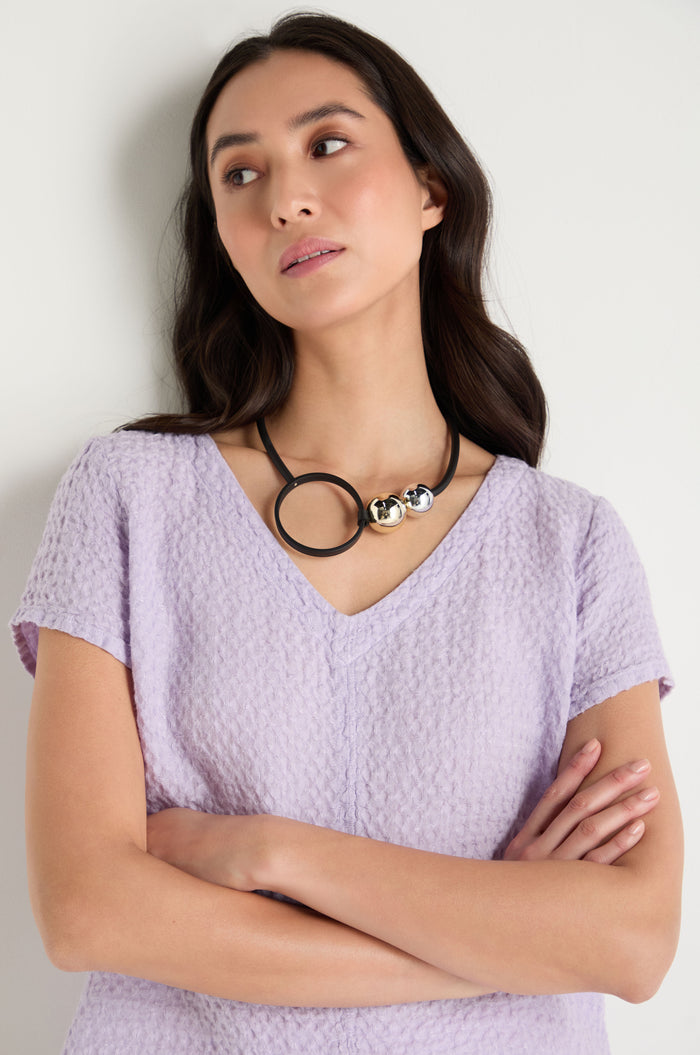 A woman with long dark hair, wearing a textured light purple top and the Space Necklace—an eye-catching piece with oversized acrylic beads—stands with arms crossed and gazes slightly to the side.