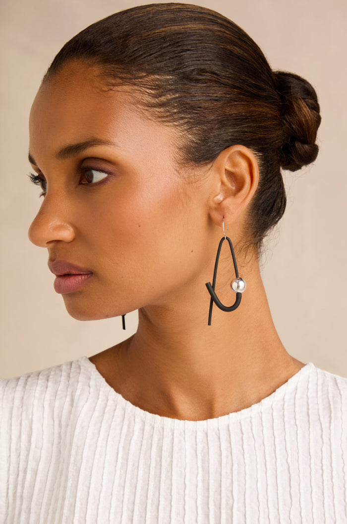 Woman with hair in a bun wearing a white textured top and the Sphere Earrings—large, abstract black contemporary earrings featuring a silver bead—shown in profile against a neutral background.