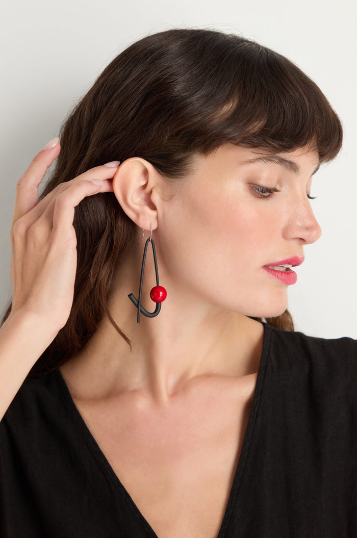 A woman in a black top wears Sphere Earrings—large geometric earrings with a red bead. Handmade in Paris, these sculptural earrings add an artistic touch to her elegant style as she looks to the side, hand near her ear.