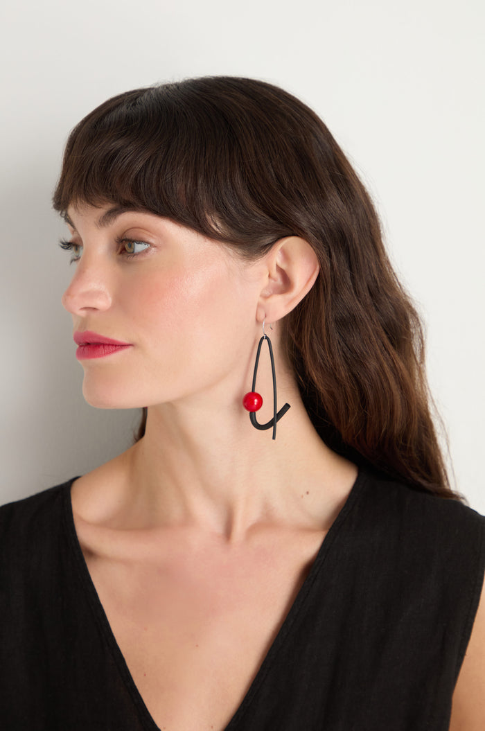 A woman with dark brown hair wears a sleeveless black top and Sphere Earrings—large black earrings with a red bead, handmade in Paris—standing against a plain light background.