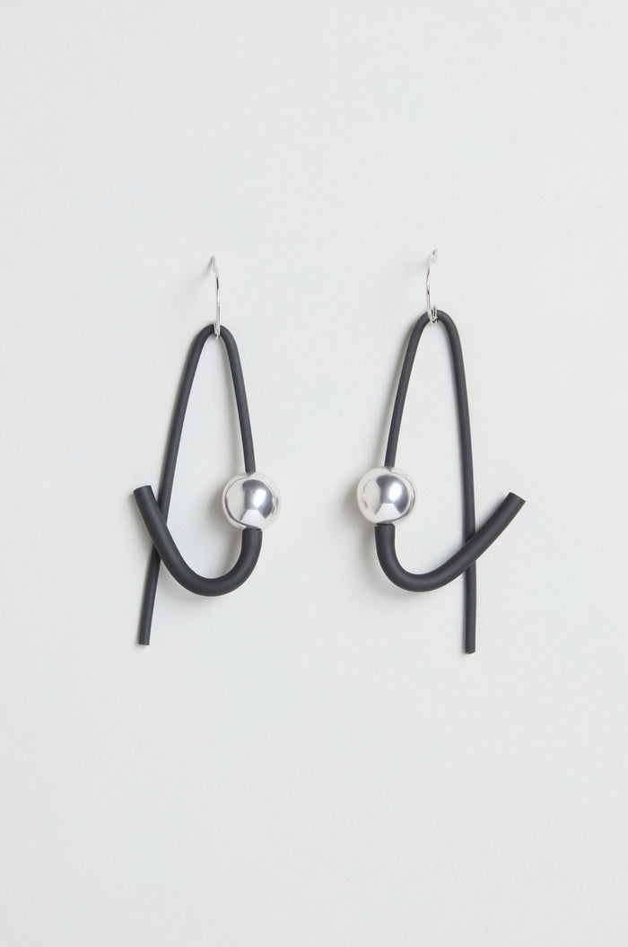 The Sphere Earrings are a pair of contemporary, handmade earrings featuring black curved loops and metallic silver spheres. Crafted in Paris, they hang from simple hooks and are displayed against a plain white background.