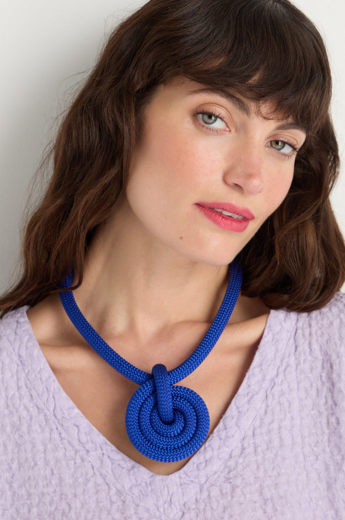 Woman with wavy brown hair wears a textured lavender top and the Spiral Cord Necklace—a bold blue handmade accessory with a spiral design, crafted in Paris—standing against a plain light background.