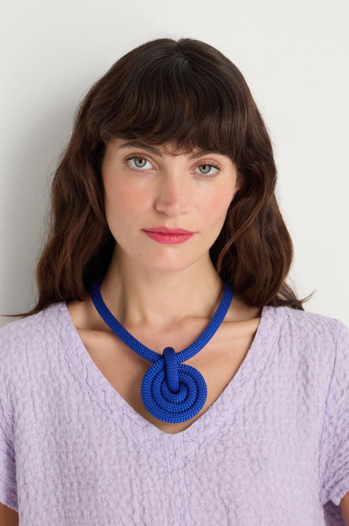 A woman with long brown hair wears a textured lavender top and the Spiral Cord Necklace, a bold blue piece handmade in Paris, standing against a plain white background.