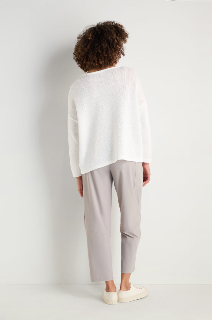 A person stands facing a white wall, wearing the Spot Cotton Knit sweater in organic cotton, paired with light gray pants and white shoes.