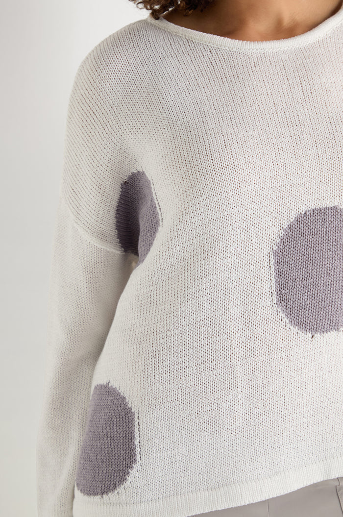 A person wears the Spot Cotton Knit, an organic cotton white sweater with a bold spot pattern; upper body partially shown against a plain background.