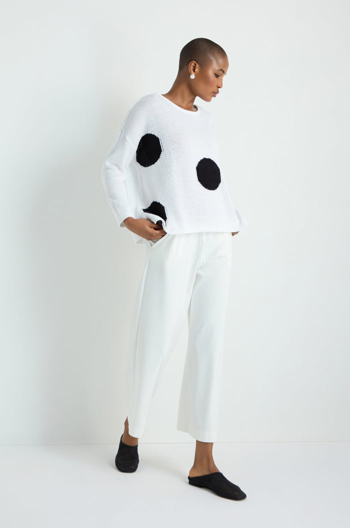 Wearing the Spot Cotton Knit sweater with bold spots, a person with long, straight, light hair pairs it with black wide-leg pants and white sneakers, standing against a plain white background.