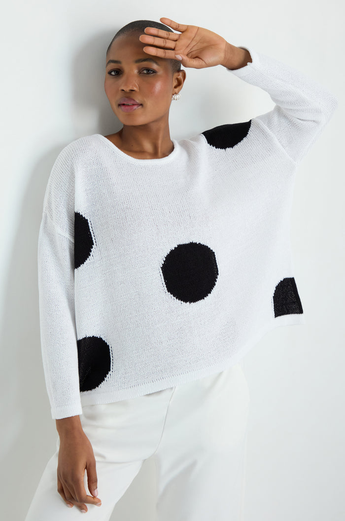 A woman with straight, long gray hair wears the Spot Cotton Knit—a white sweater with a bold spot pattern made from organic cotton—paired with black pants, standing against a plain white background with her hands in her pockets.
