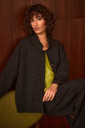 A person with short curly hair sits against a dark wooden wall, wearing the Square Jacquard Jacket—a dark-patterned, cotton-rich piece—layered over a bright green top.