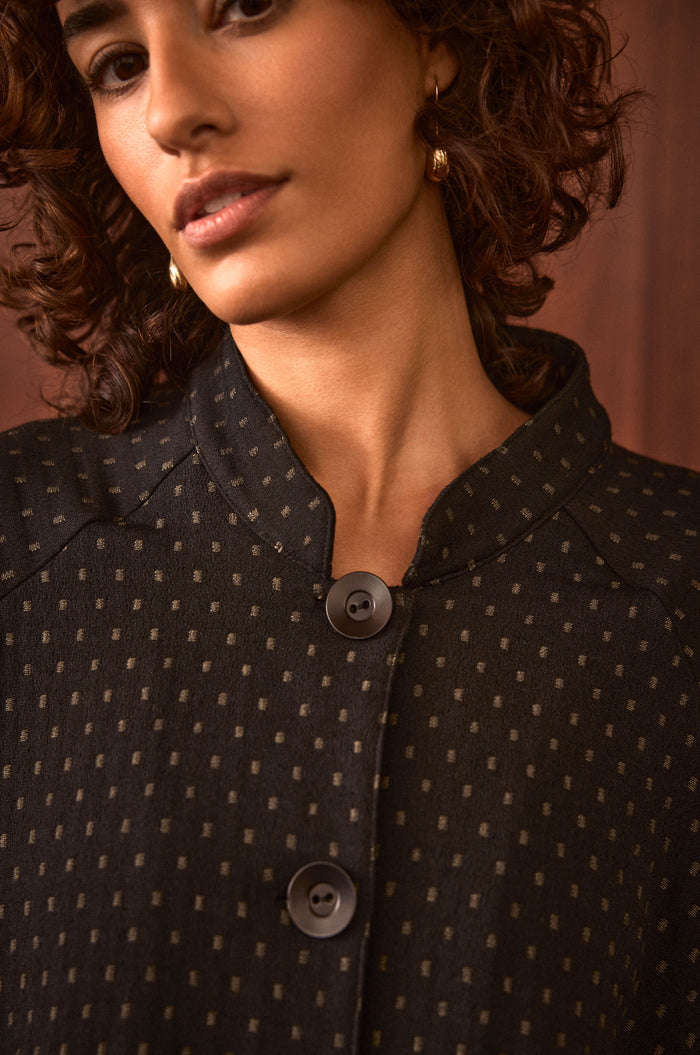 A woman stands against a brown background wearing the Square Jacquard Jacket—a black, cotton-rich jacket with a subtle square pattern and two large buttons.