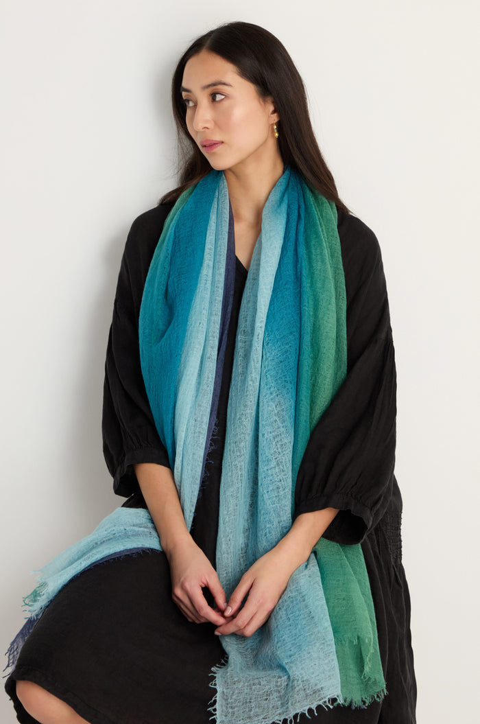 A woman with long dark hair, in a dark dress, wears the St. Tropez Cashmere Shawl—a large blue and green gradient wrap—while seated against a plain light-colored background.