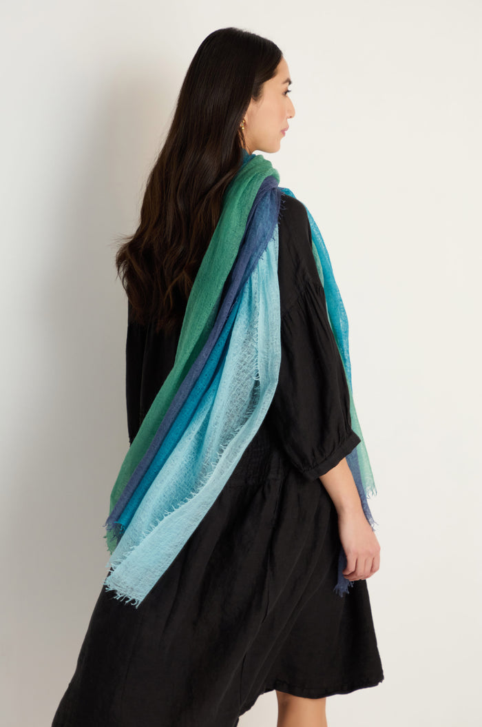 A woman in a black dress stands sideways, elegantly draping the St. Tropez Cashmere Shawl in green and blue tones—ideal for lightweight layering—against a plain white background, capturing effortless style.