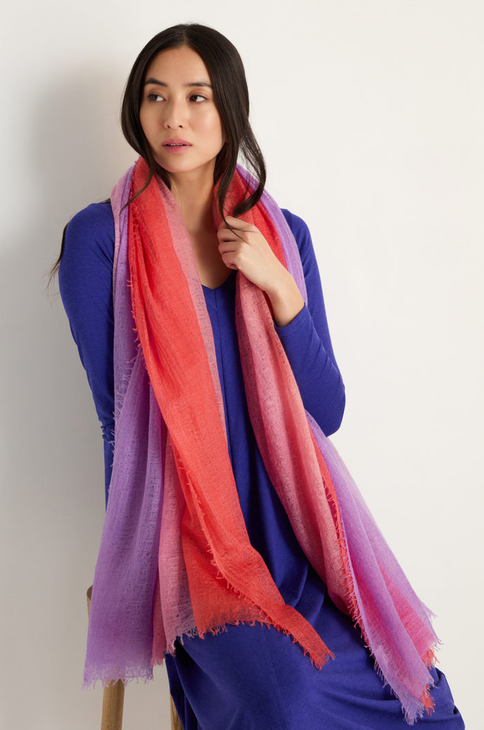 Woman in a long-sleeved blue dress and the St. Tropez Cashmere Shawl, featuring ombre red, pink, and purple hues, looks to the side against a plain light background—ideal for lightweight layering in St. Tropez.