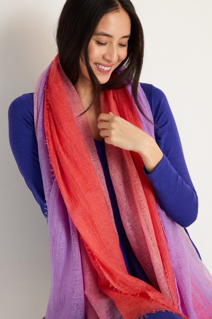 A woman in a blue top wears the St. Tropez Cashmere Shawl—large, lightweight, and featuring red, pink, and purple gradient tones—smiling as she looks down, perfect for effortless layering in St. Tropez.