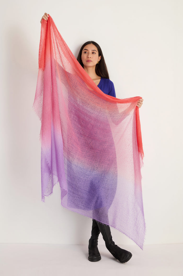 A woman stands against a plain background, holding up the St. Tropez Cashmere Shawl—a large, semi-transparent accessory with red, pink, and purple gradients—ideal for lightweight layering on breezy St Tropez evenings.