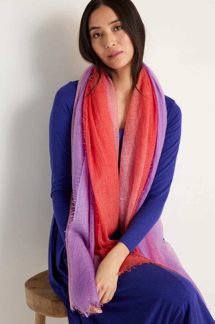A woman in a long-sleeve blue dress sits on a stool, wearing the St. Tropez Cashmere Shawl in red and purple for lightweight layering, looking directly at the camera.