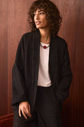 A person with curly hair is wearing the Stitched Jacquard Jacket, which features a quilted pattern, paired with a white shirt and a necklace adorned with a red bead, standing in front of a wooden wall.