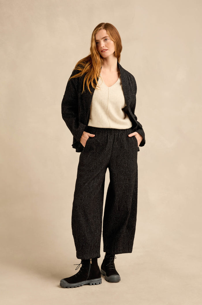 A woman stands with her hands in her pockets, wearing a cream V-neck top, the Stitched Jacquard Jacket with a boxy silhouette, matching wide-leg pants, and black boots against a beige background.